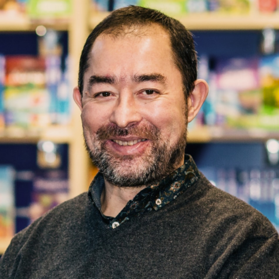 A man with a short beard and wearing a dark sweater, smiling, sits in front of a background filled with colorful books. The man is Martin Heng.