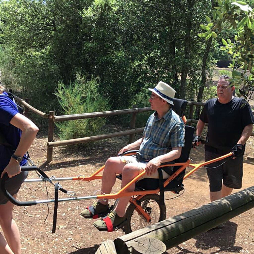 Martin Heng is seated in a Joëlette hiking wheelchair, being assisted by two people on a wooded trail. One person is pulling from the front, while the other is pushing from behind. Martin is wearing a hat, a plaid shirt, and shorts.