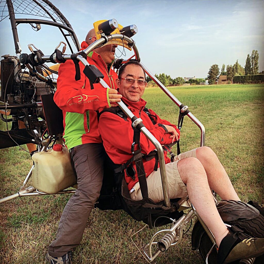 Martin Heng is seated in a powered paraglider with another person behind him, both wearing red jackets. They are on a grassy field, ready for takeoff, with the paraglider's equipment visible behind them. Martin is smiling, looking content and excited.