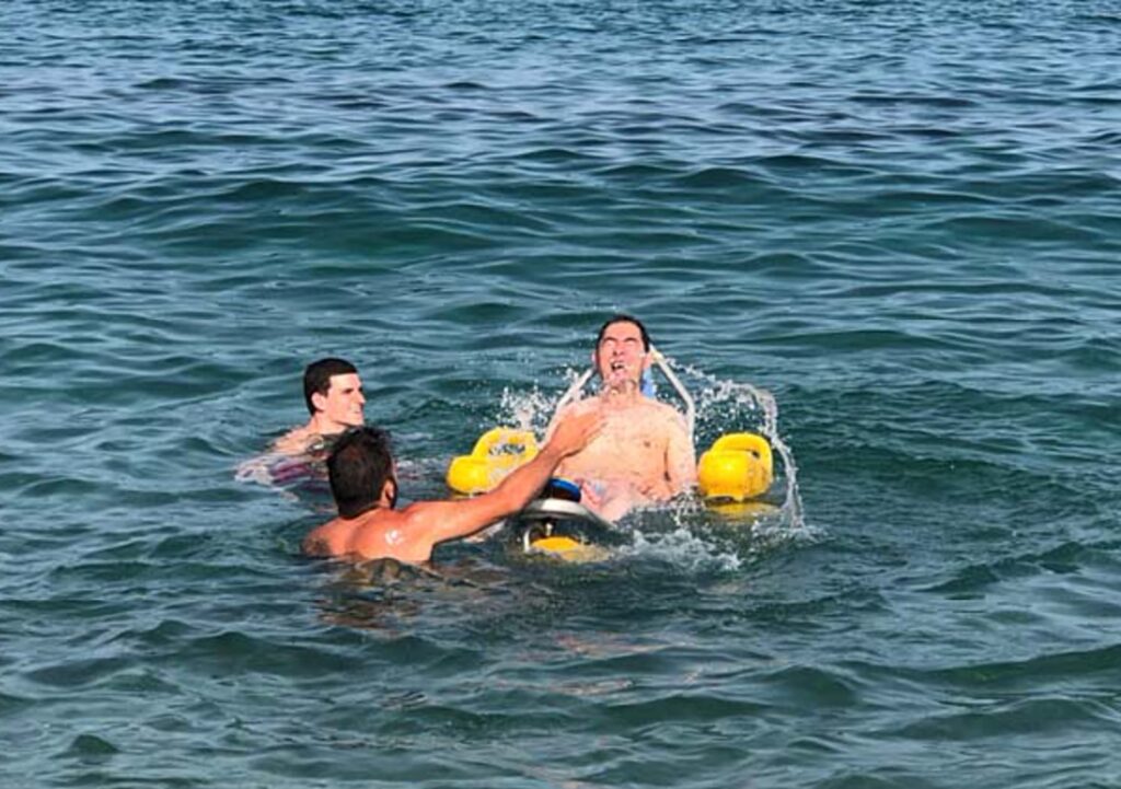 Martin Heng is floating in the sea on a water wheelchair with yellow floats. Two other people are in the water nearby, assisting him. Martin appears to be enjoying the experience, with a splash of water around him.