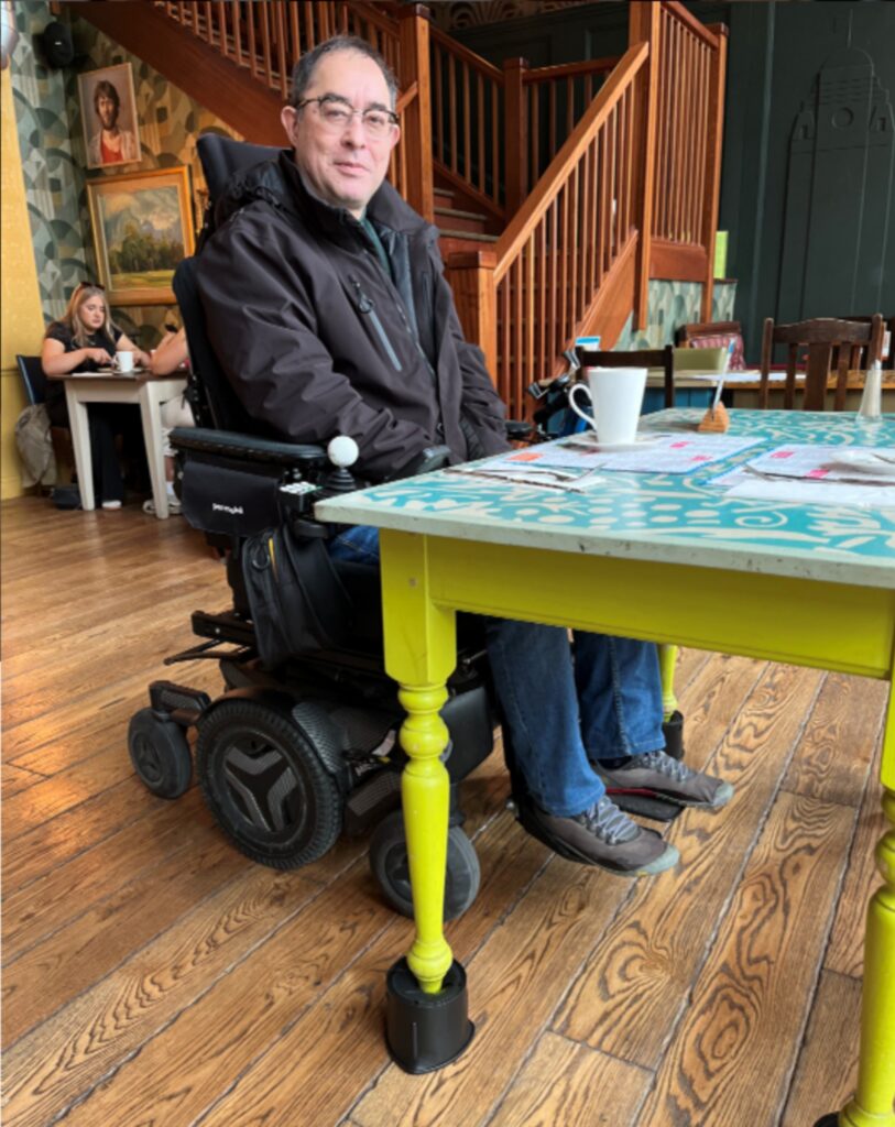 Martin Heng is seated in a motorized wheelchair at a table in a cafe. He is wearing a dark jacket and glasses, smiling slightly. The table has bright yellow legs and is raised on black blocks to accommodate his wheelchair. The cafe has wooden floors, and other patrons are visible in the background.